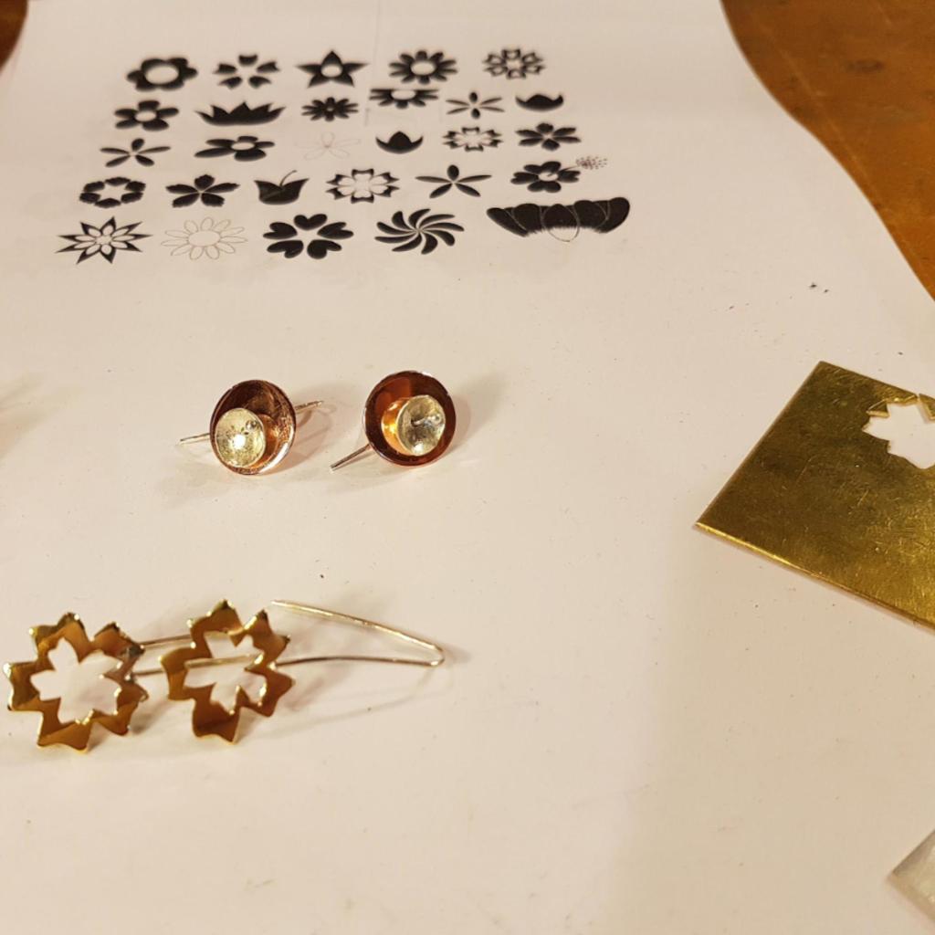 A flat lay image featuring two pairs of earrings: one pair with copper and silver elements and another with a floral design, alongside sheets with various flower illustrations.
