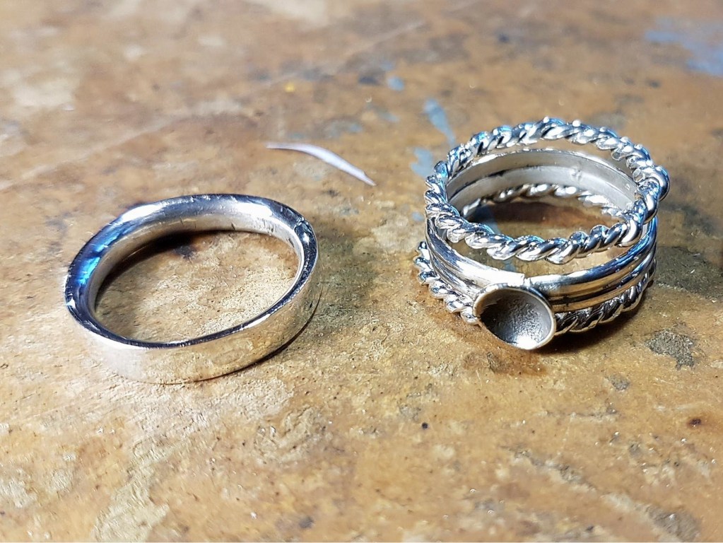 A smooth silver wedding band next to a stack of twisted silver rings featuring a small funnel design.